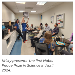 A picture of Kristy holding up a gift bag in a crowded breakroom while 
              everyone looks on in entertained curiosity.